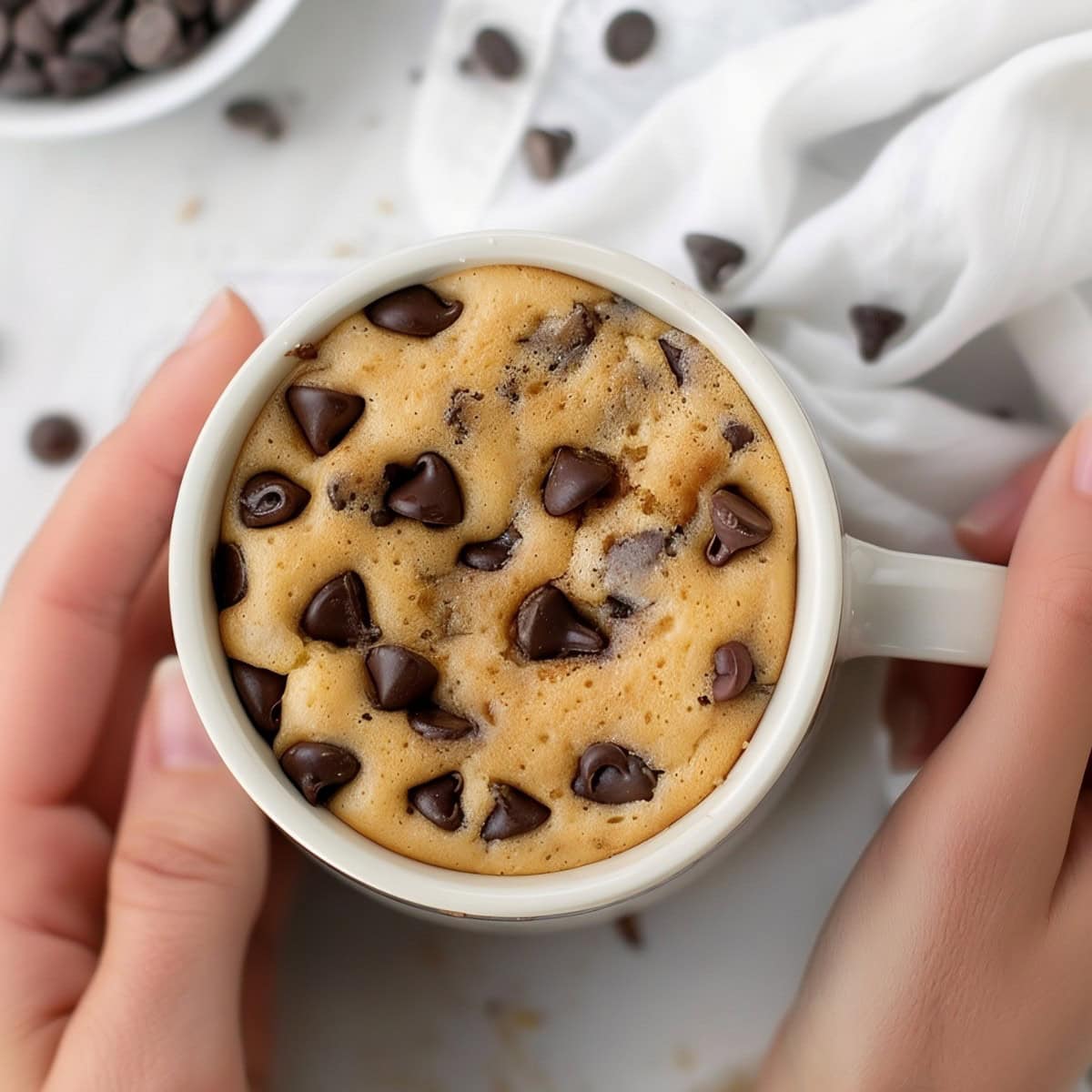 A hand holding a chocolate chip mug cake, top down view