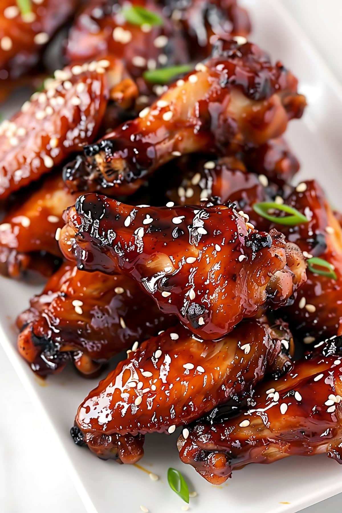 Bunch of honey glazed chicken wings garnished with sesame seeds and chopped green onions in a white plate.