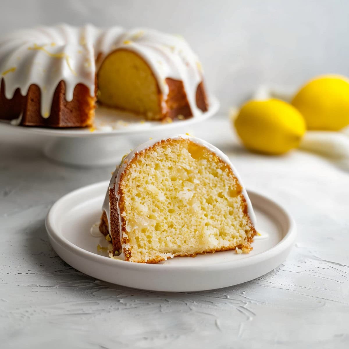 A piece of lemon bundt cake on a plate with the rest of the cake behind on a cake stand