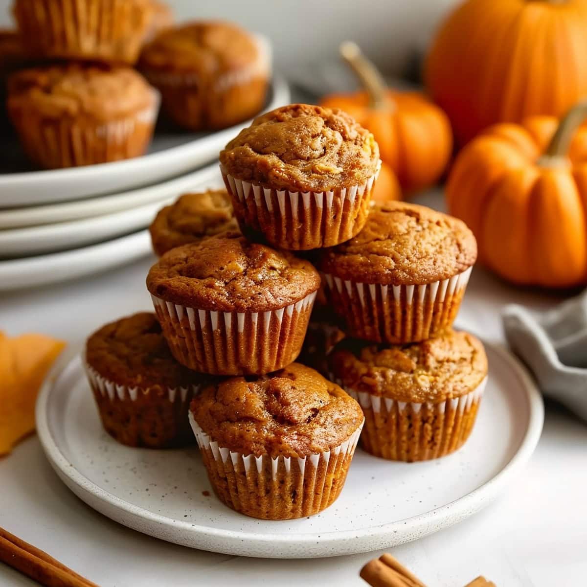 Pumpkin banana muffins with cinnamon stacked on a white plate with pumpkins in the background.