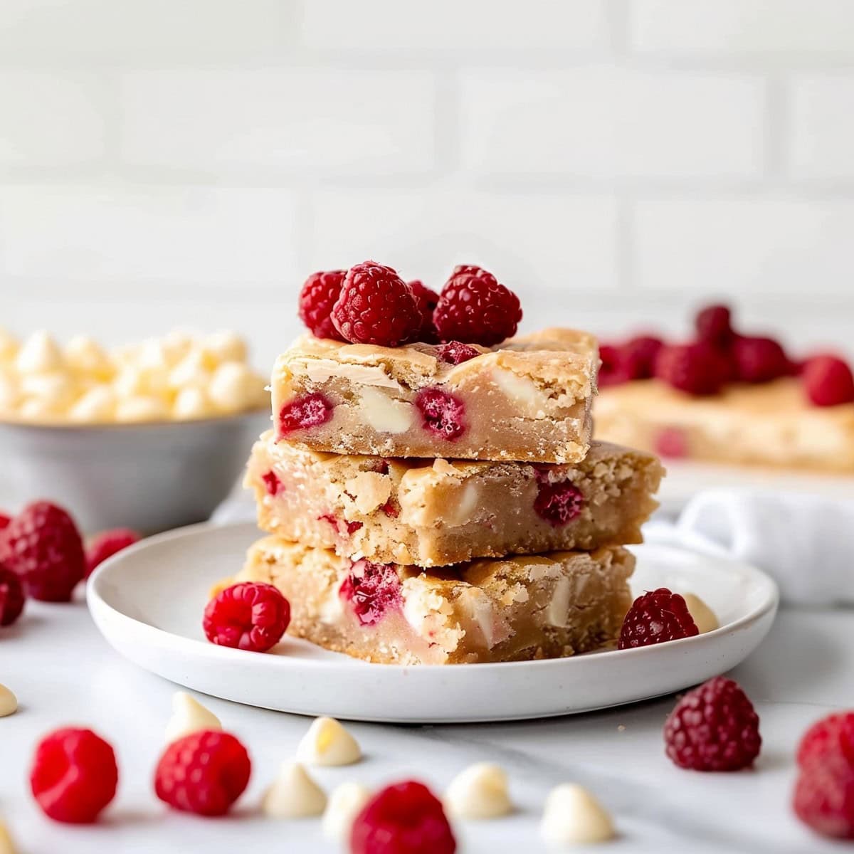 Raspberry and White Chocolate Blondies Stacked in a White Plate with white chocolate chips and fresh berries around