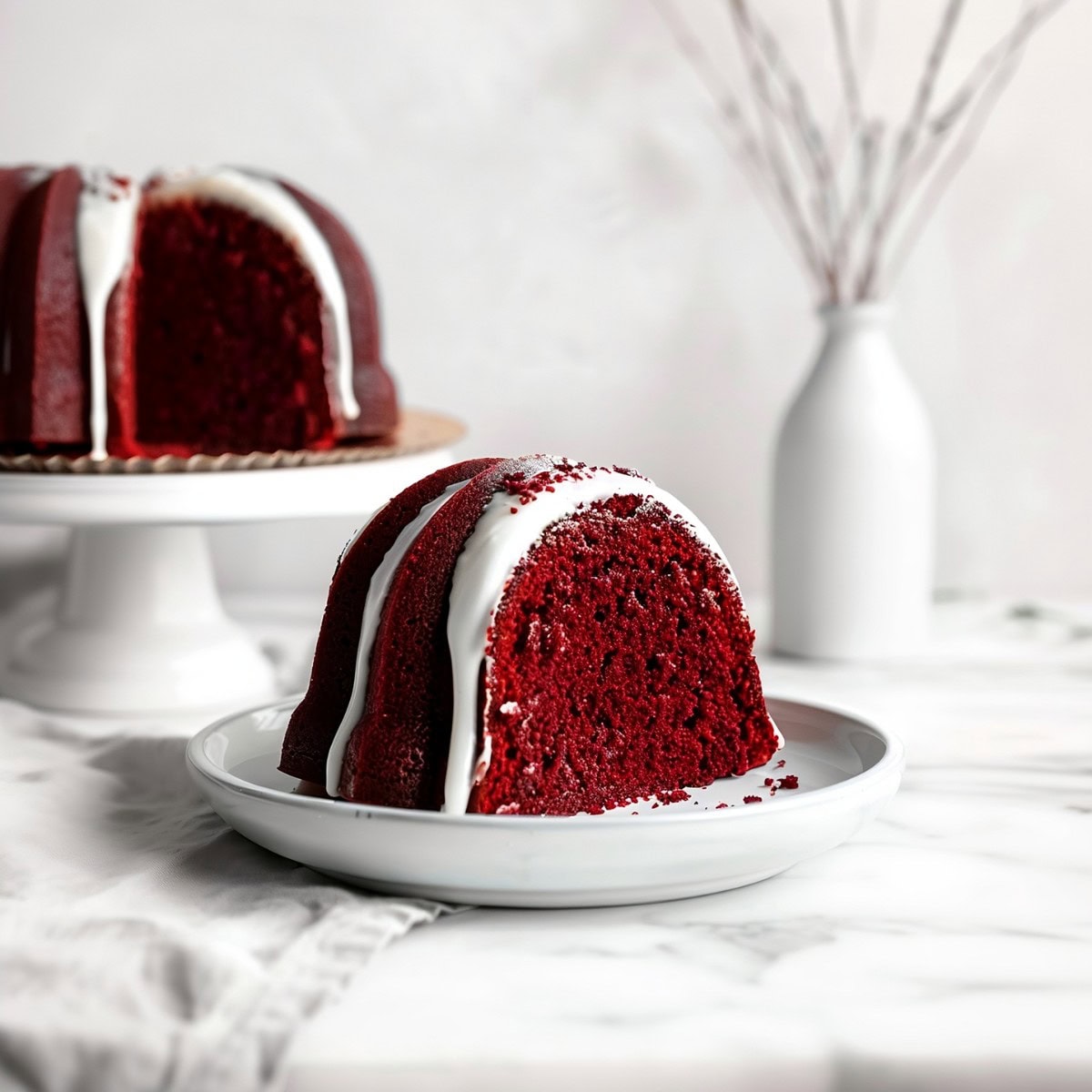 A piece of red velvet bundt cake on a plate with the whole cake in the background on a cake stand