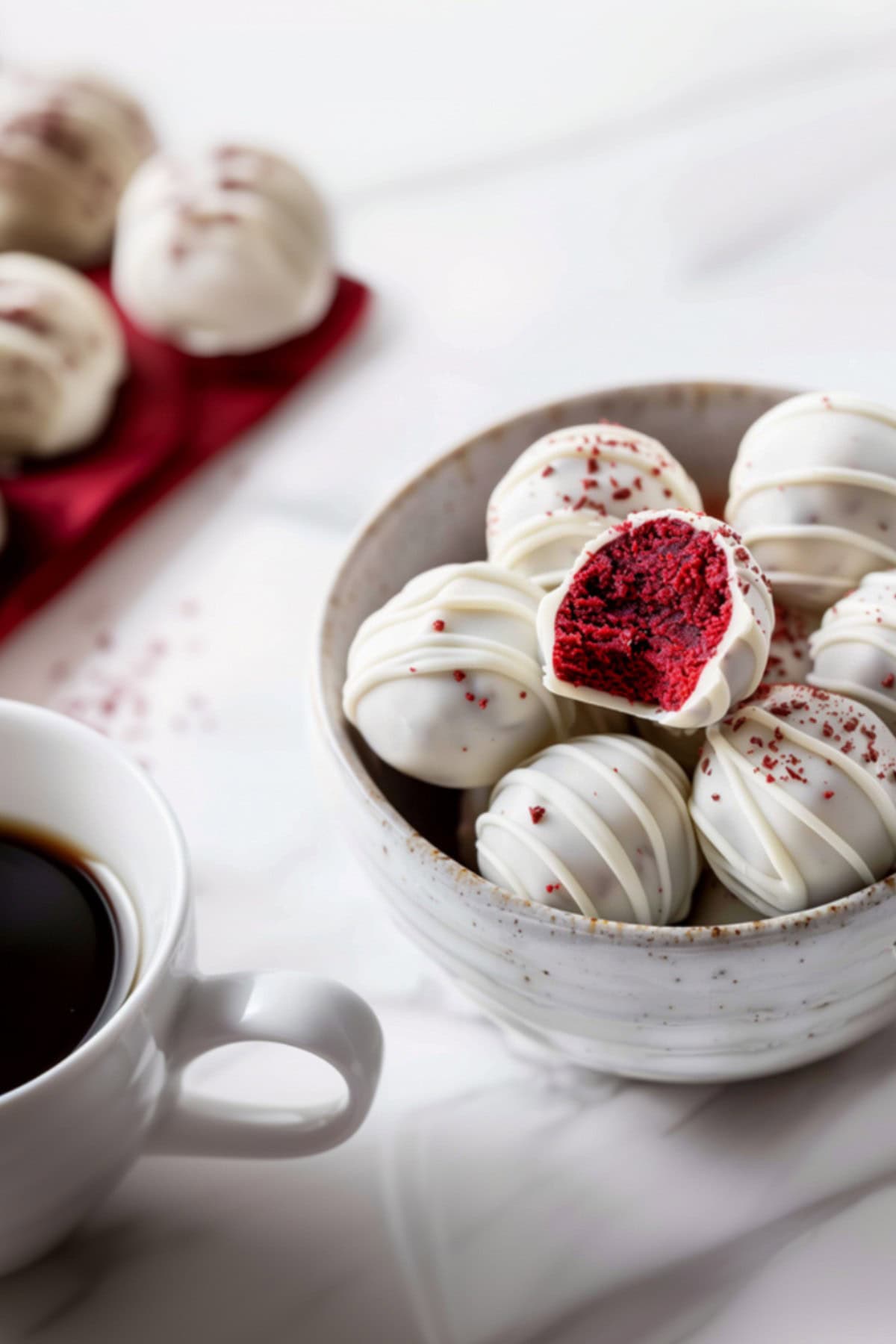 A red velvet truffle with a bite taken out, revealing its rich, velvety interior, in a bowl of red velvet truffles next to a cup of coffee