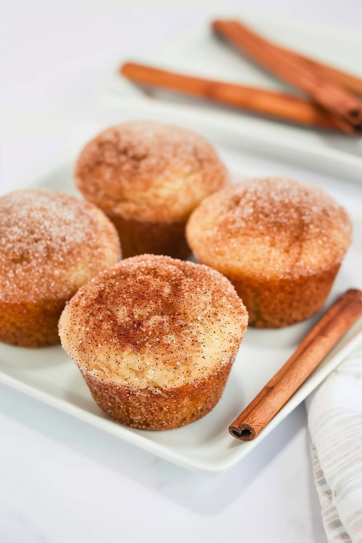 Four Snickerdoodle muffins on a white plate with a cinnamon stick on the side