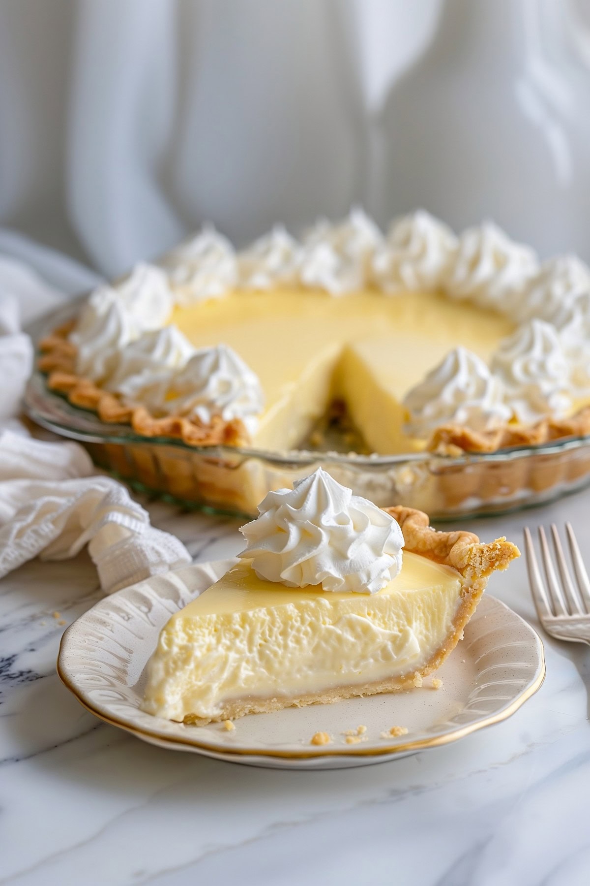 A slice of vanilla cream pie in a white plate, whole pie dish with slice missing in the background.