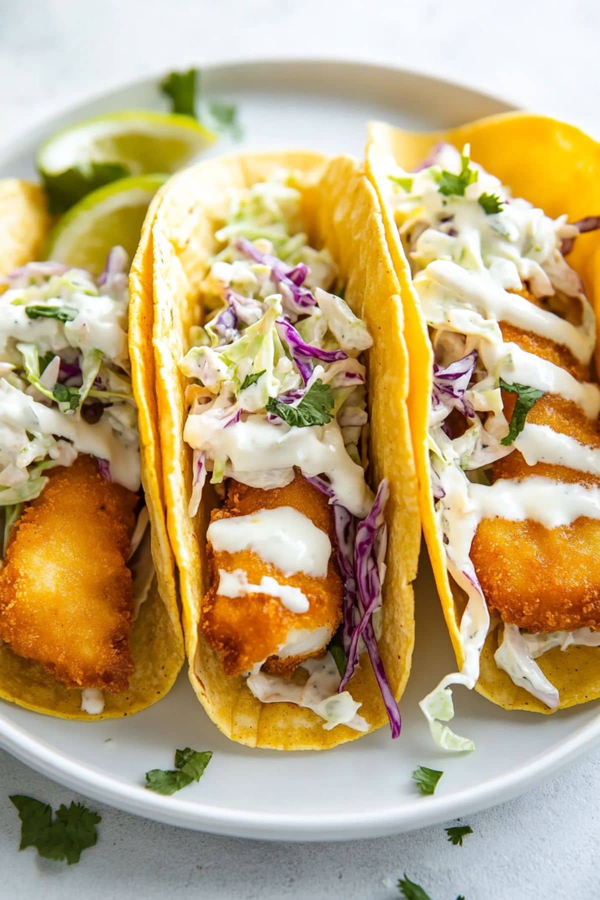 Crispy Baja-style beer battered fish tacos drizzled with white sauce over a coleslaw served on a white plate.