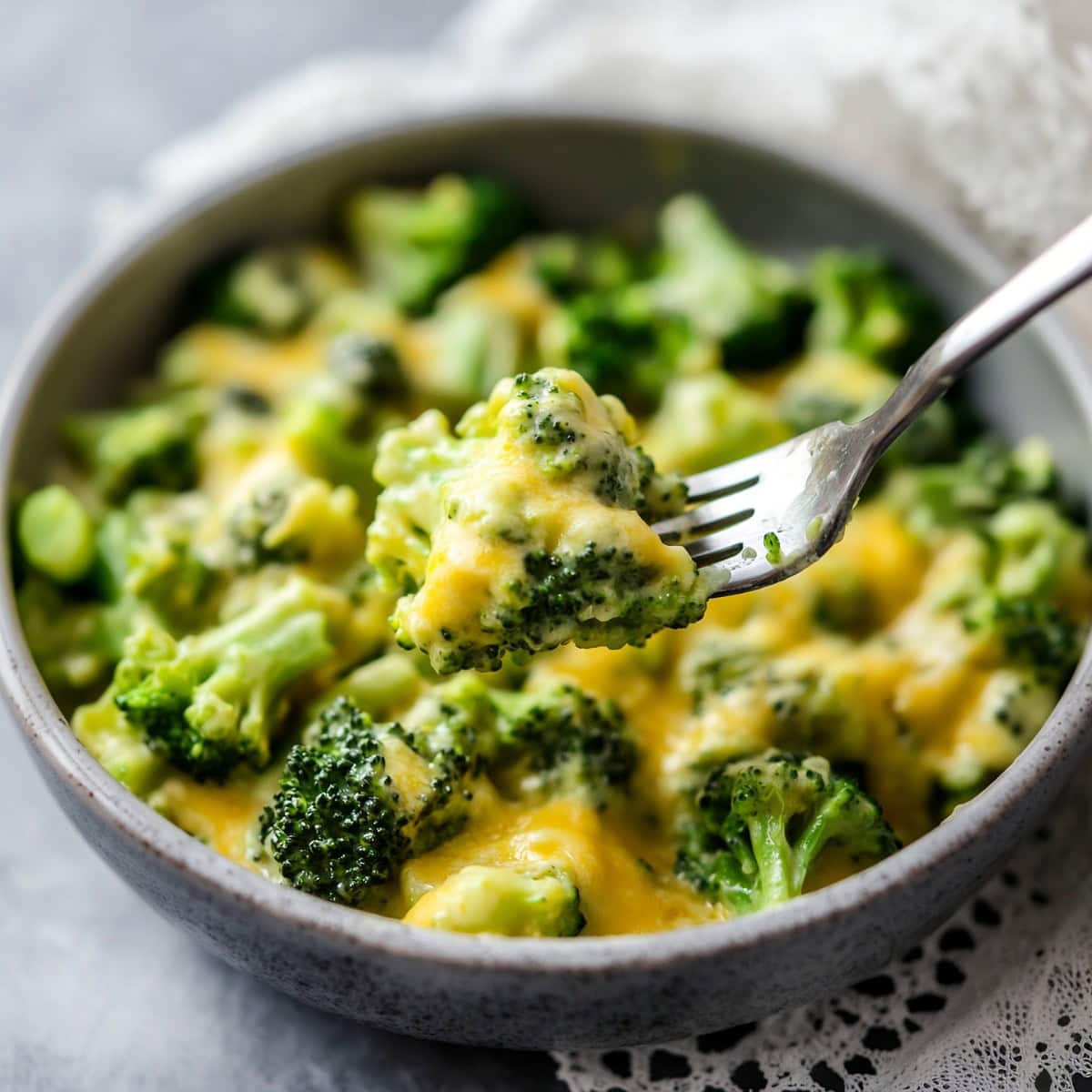 A fork lifting a piece of cheesy broccoli from a bowl.