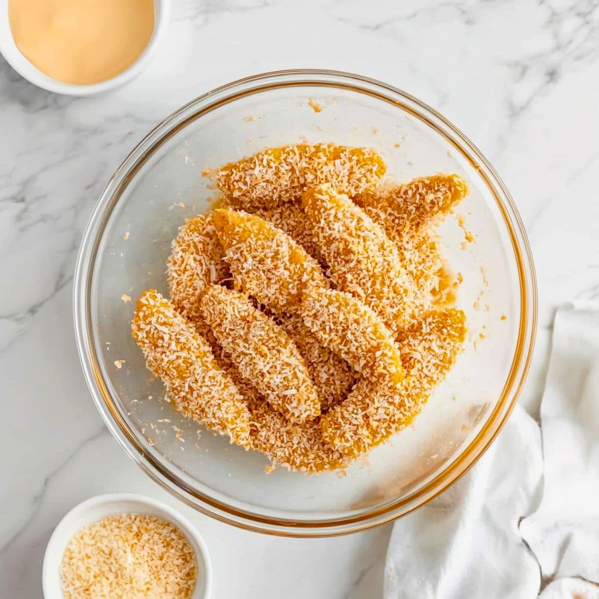 Chicken tenders coated in bread crumbs and sweetened coconut flakes mixture in a glass mixing bowl.