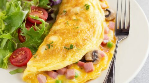 A close-up of Denver omelette with mushrooms, ham bits and bell peppers with vegetable salad and fork on the side in a plate