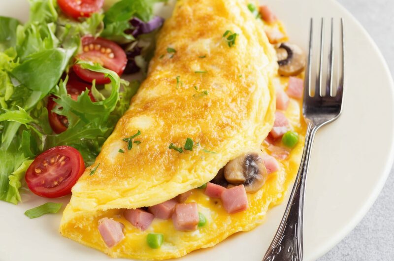 A close-up of Denver omelette with mushrooms, ham bits and bell peppers with vegetable salad and fork on the side in a plate