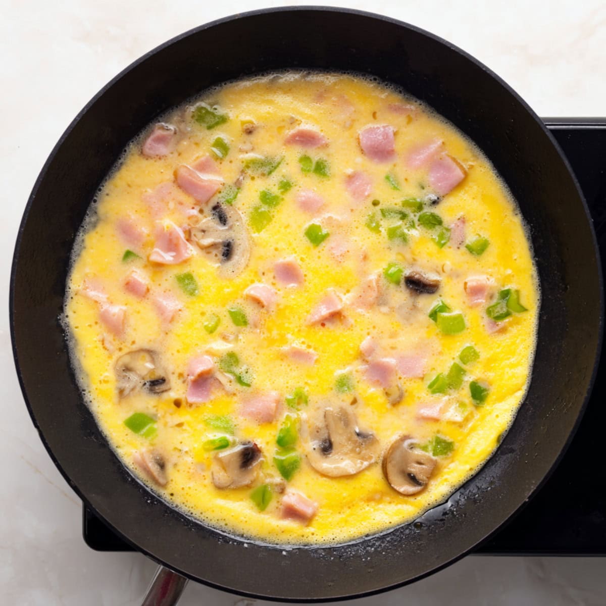 Denver omelette cooking in a black skillet, with visible pieces of diced ham, green bell peppers, and sliced mushrooms in the egg mixture