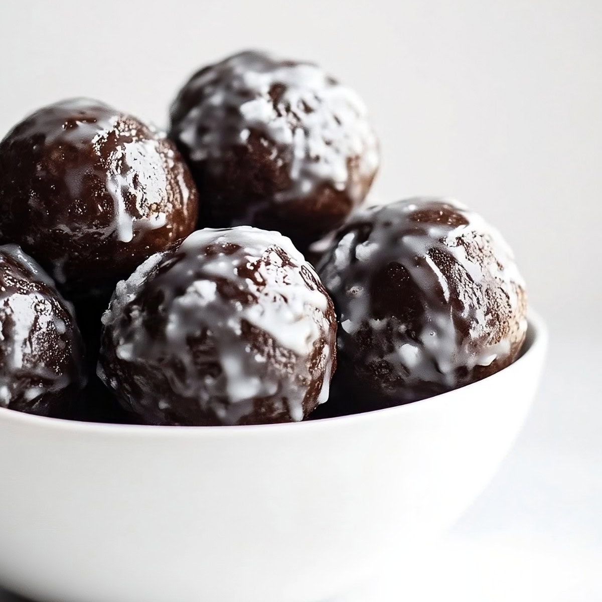 A close-up shot of glazed chocolate donut holes in a white bowl, side view