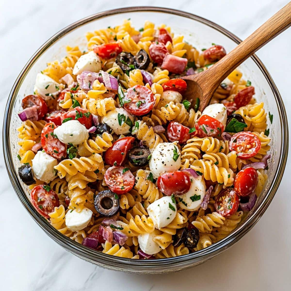 Italian pasta salad with rotini pasta, olives, mozzarella balls, tomatoes and salami tossed in a glass bowl.