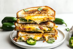 Jalapeno popper grilled cheese sandwich with melted cheddar cheese, crumbled bacon and roasted jalapeno filling served on a white plate.