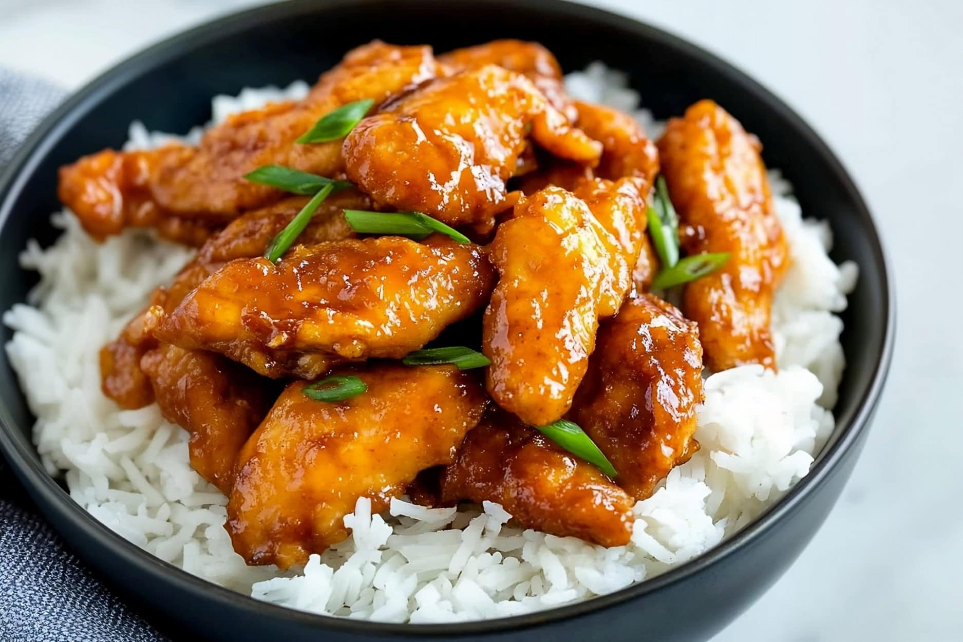 Succulent Mongolian chicken served on a black ceramic bowl on a white marble table.