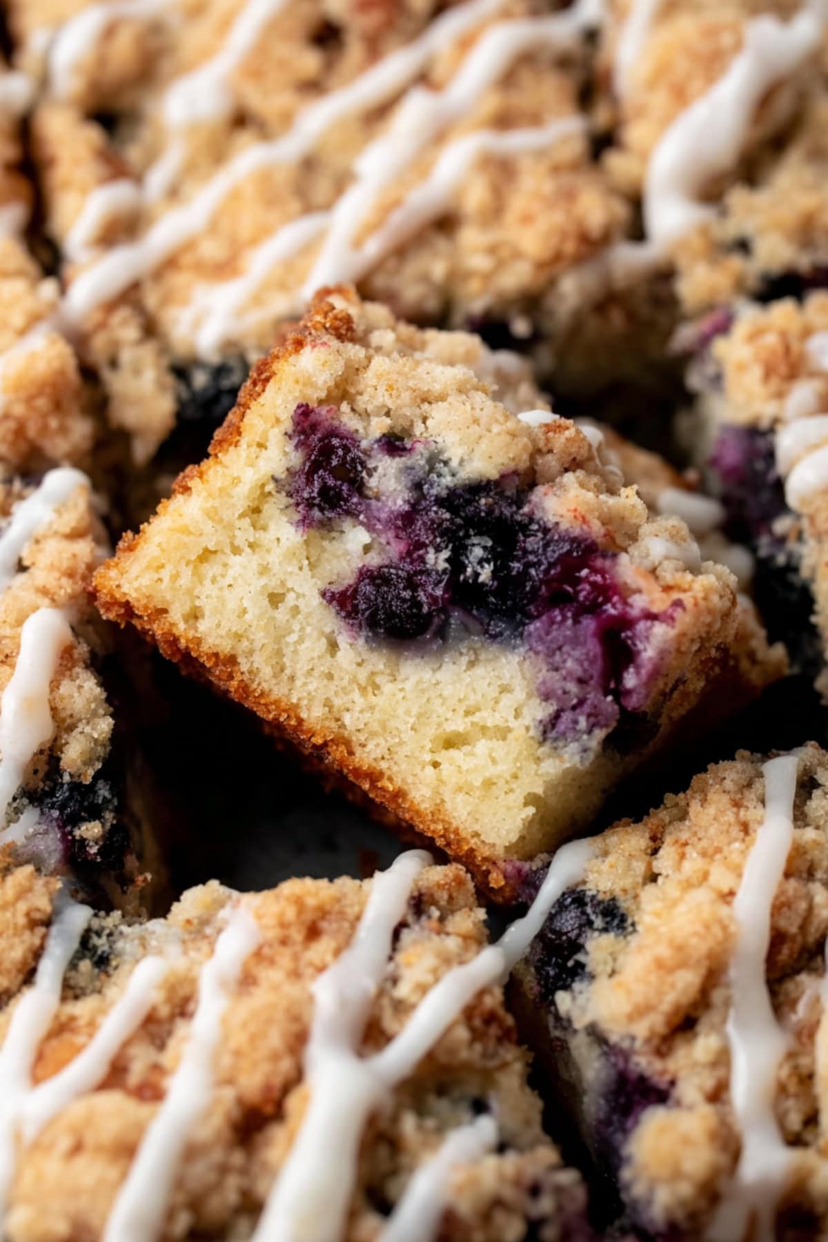 A close-up of a blueberry crumb cake slice with a golden crumb, juicy blueberries, and a drizzle of white glaze on top.