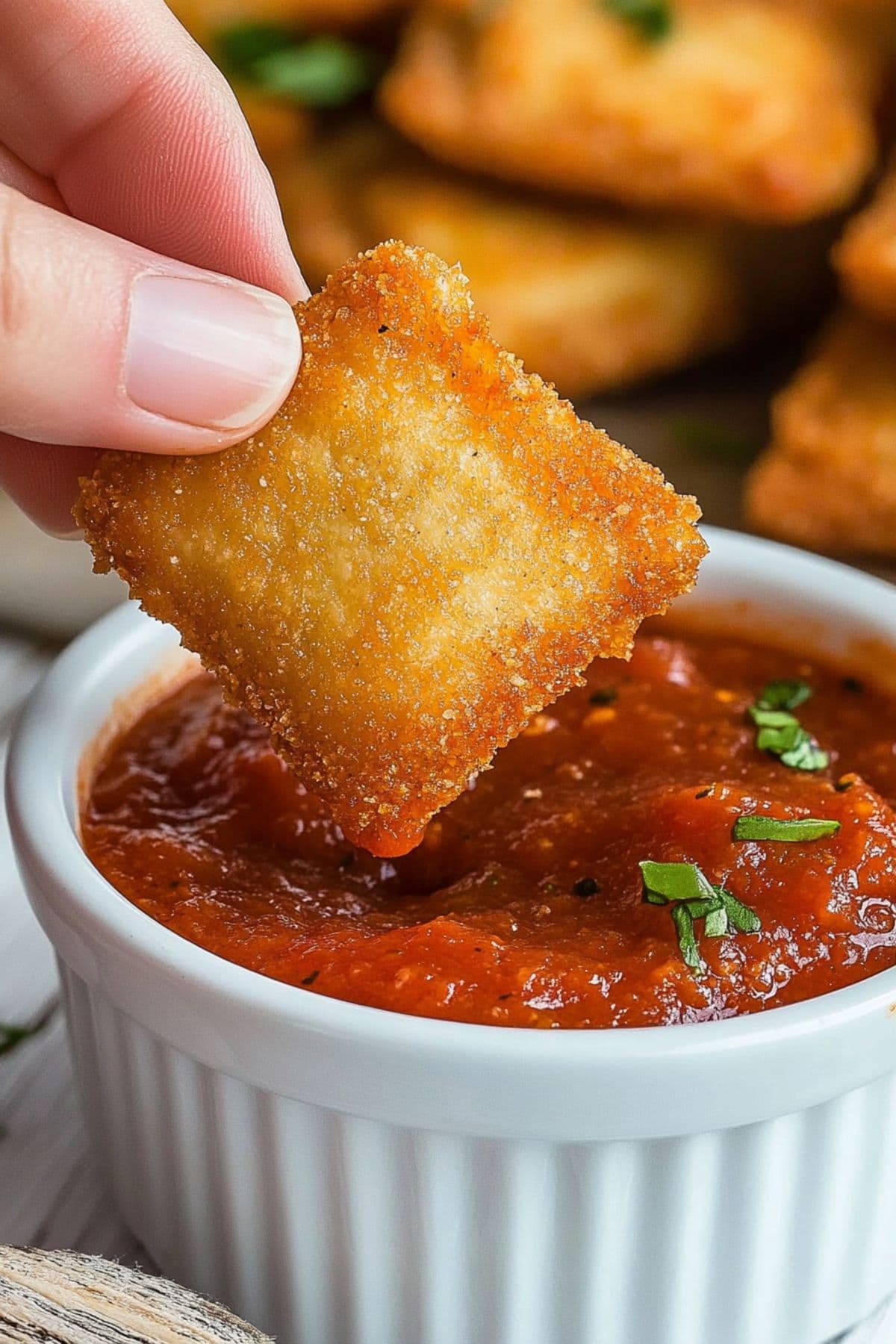 Hand holding a piece of fried breaded ravioli dipped in marinara on a white wooden table, close up.