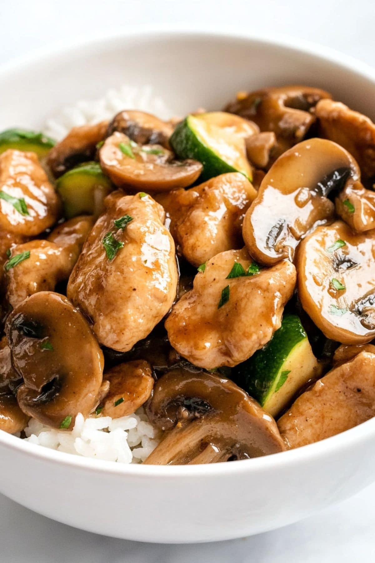 Chicken with mushroom and sliced zucchini tossed in sauce served on top of a white rice in a white bowl.