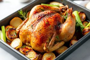 Roasted whole chicken in a roasting pan with onions, garlic and celery stalks.