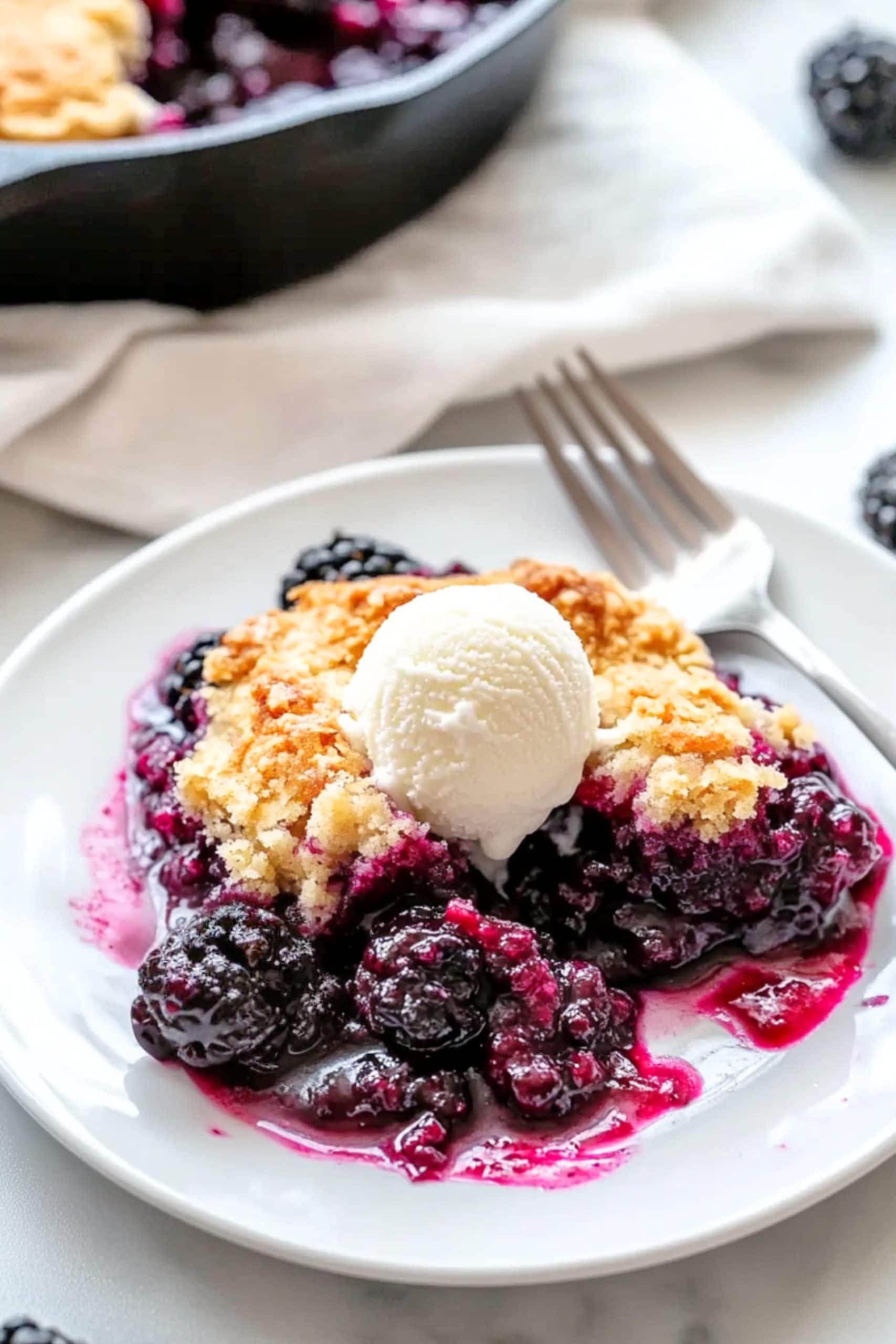 Serving of blackberry cobbler in a white plate with vanilla ice cream. 