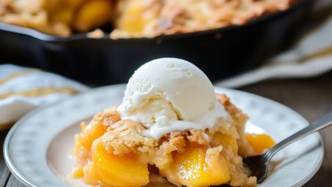 Serving of peach cobbler in a white plate with scoop of vanilla ice cream on top.