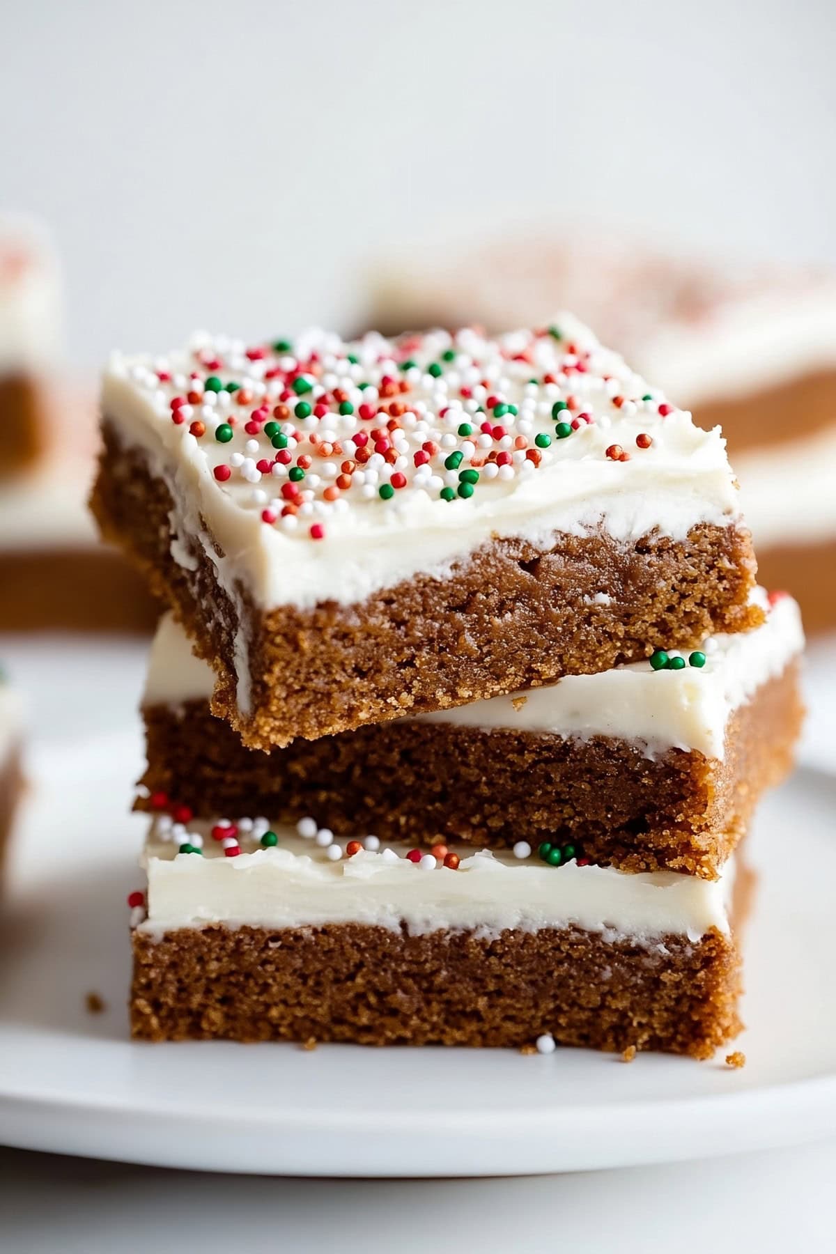 Gingerbread bars with cream cheese frosting and candy sprinkles stacked on a white plate, side view