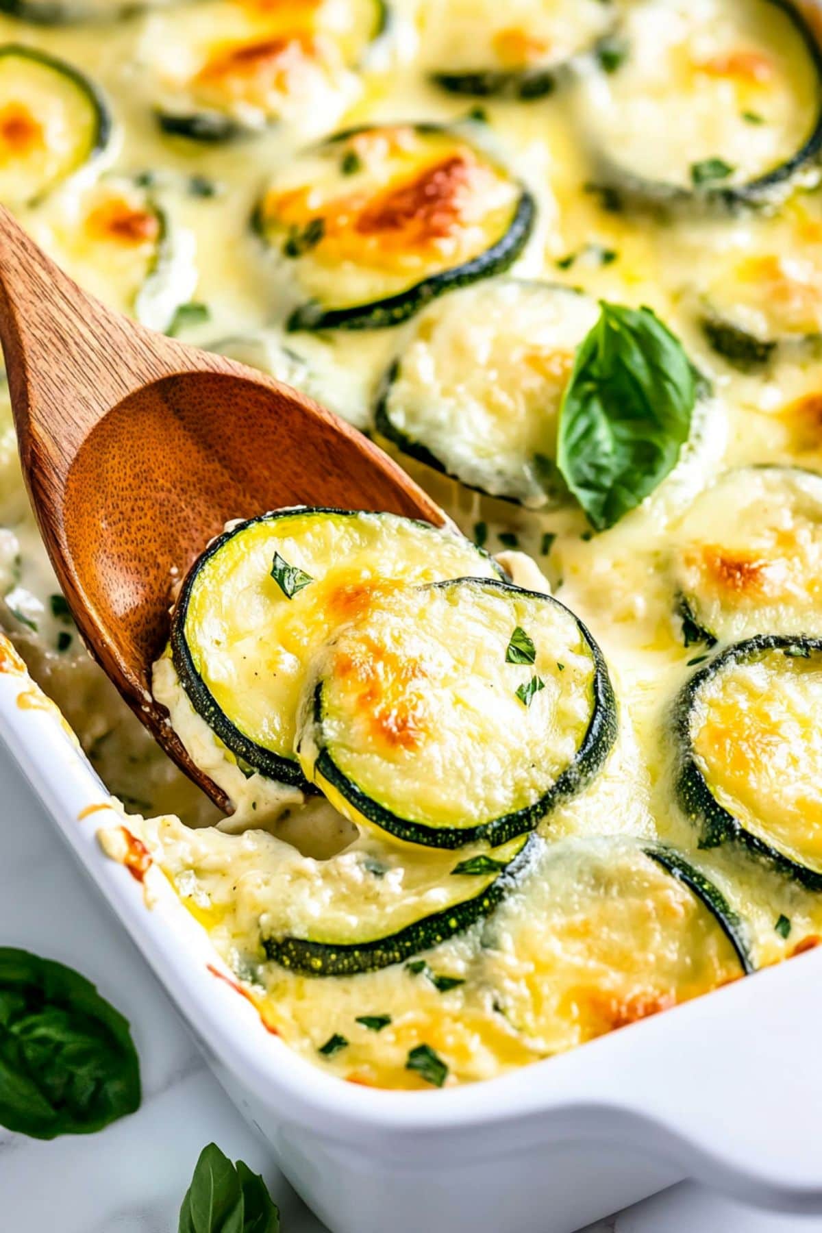 Cheesy scalloped zucchini in a white casserole dish lifted with a wooden spoon, close up.