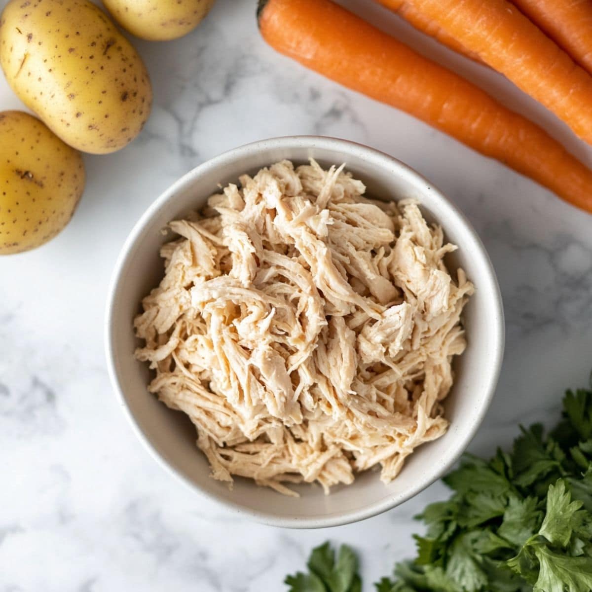 Shredded chicken in a white bowl, carrots and potatoes next to it lying on a white marble table.