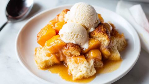 Peach cobbler with chunks of cinnamon rolls served in a white plate with vanilla ice cream.