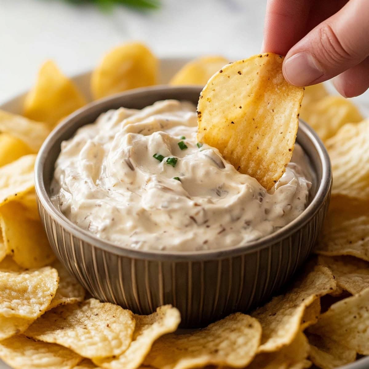 Hand holding a potato chip dipping it a bowl of French onion dip.