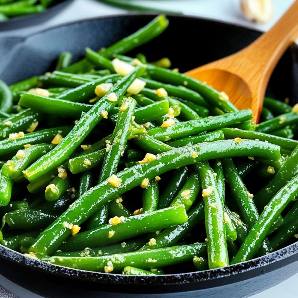 Wooden ladle tossing green beans with garlic and butter in a cast iron skillet