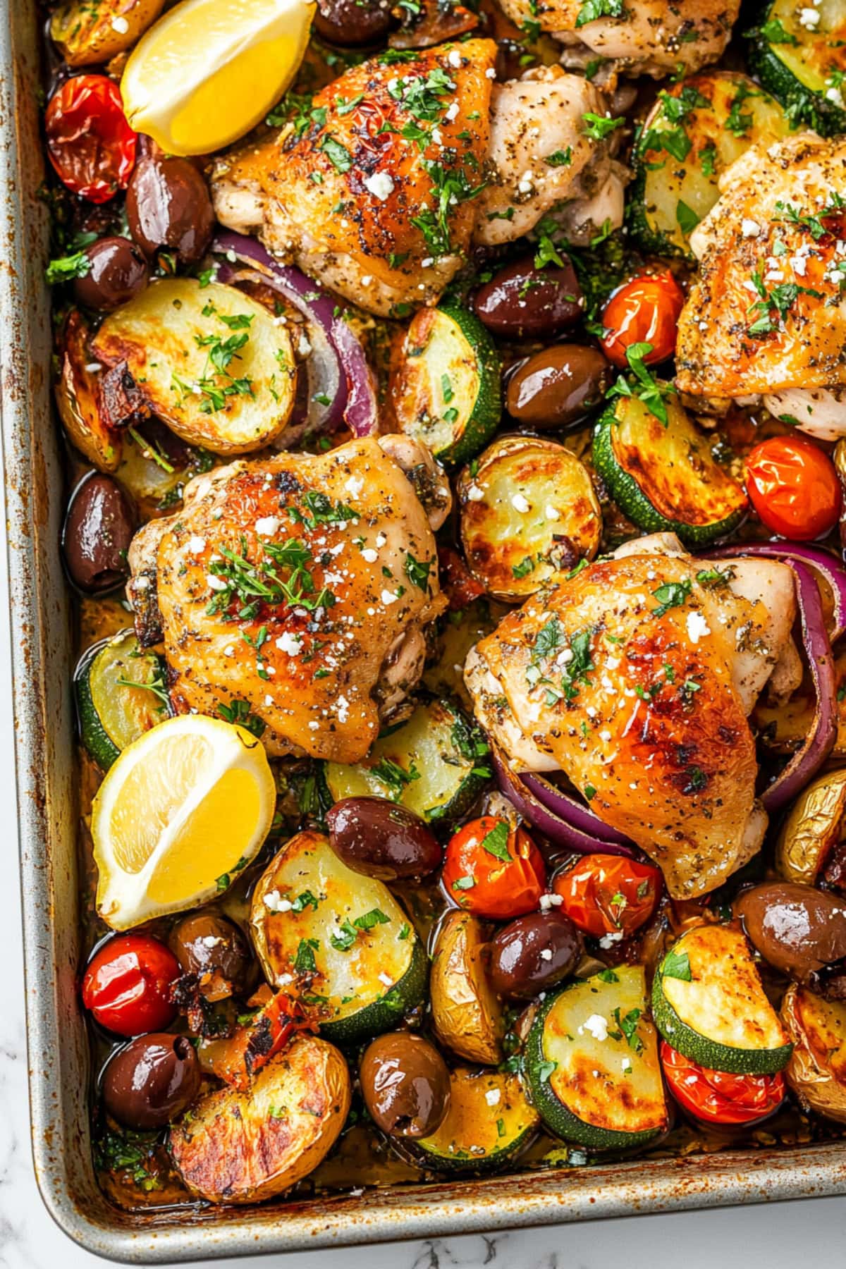 Homemade greek sheet pan chicken with colorful olives, tomatoes, cucumber, garnished with herbs.