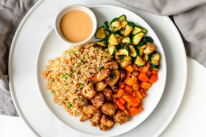 Hibachi chicken served on a white plate surrounded by colorful vegetables, fried rice and yum yum sauce.