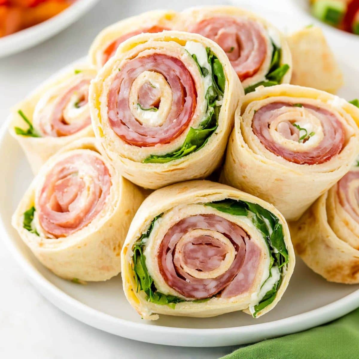 Bunch of pinwheels with lettuce, provolone cheese, salami, ham, and pepperoni provolone cheese, salami, ham, and pepperoni filling inside arranged in a white plate.