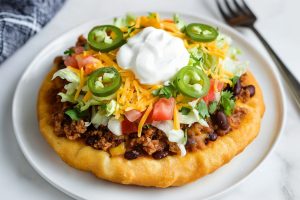 A plate of Navajo tacos with multiple toppings, including shredded lettuce, jalapeno peppers, tomatoes and shredded cheddar cheese.