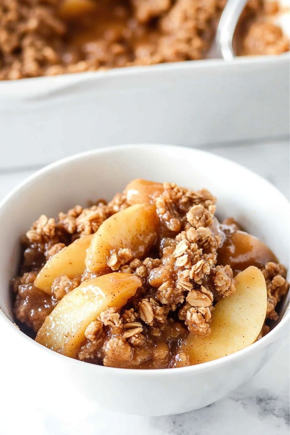 Peanut butter apple crisp served in a white bowl, rectangular baking dish with the rest of the dessert in the backfground.