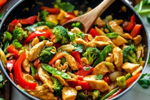 Chicken stir fried with broccoli, bell peppers, cabbage and chopped nuts in a wok pan.