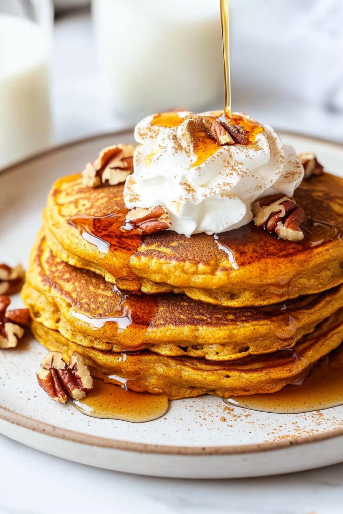 Stacked pumpkin spice pancakes with whipped cream and chopped pecans, drizzled with maple syrup.