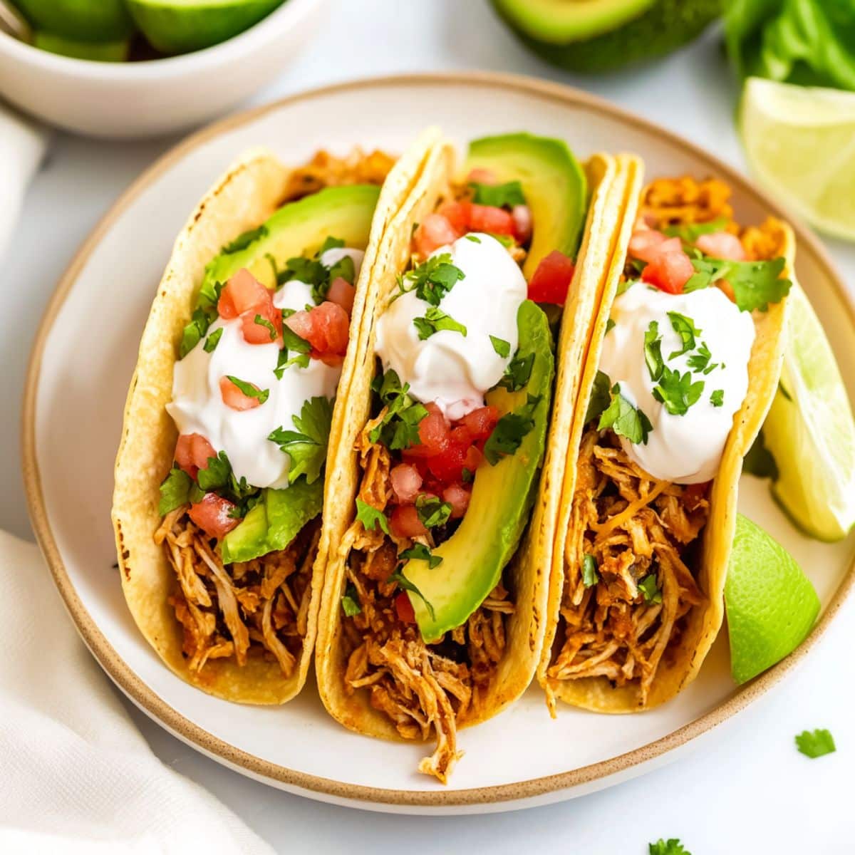 Serving of chicken tacos with sliced avocados, sour cream, chopped parsley and sour cream arranged in a plate.