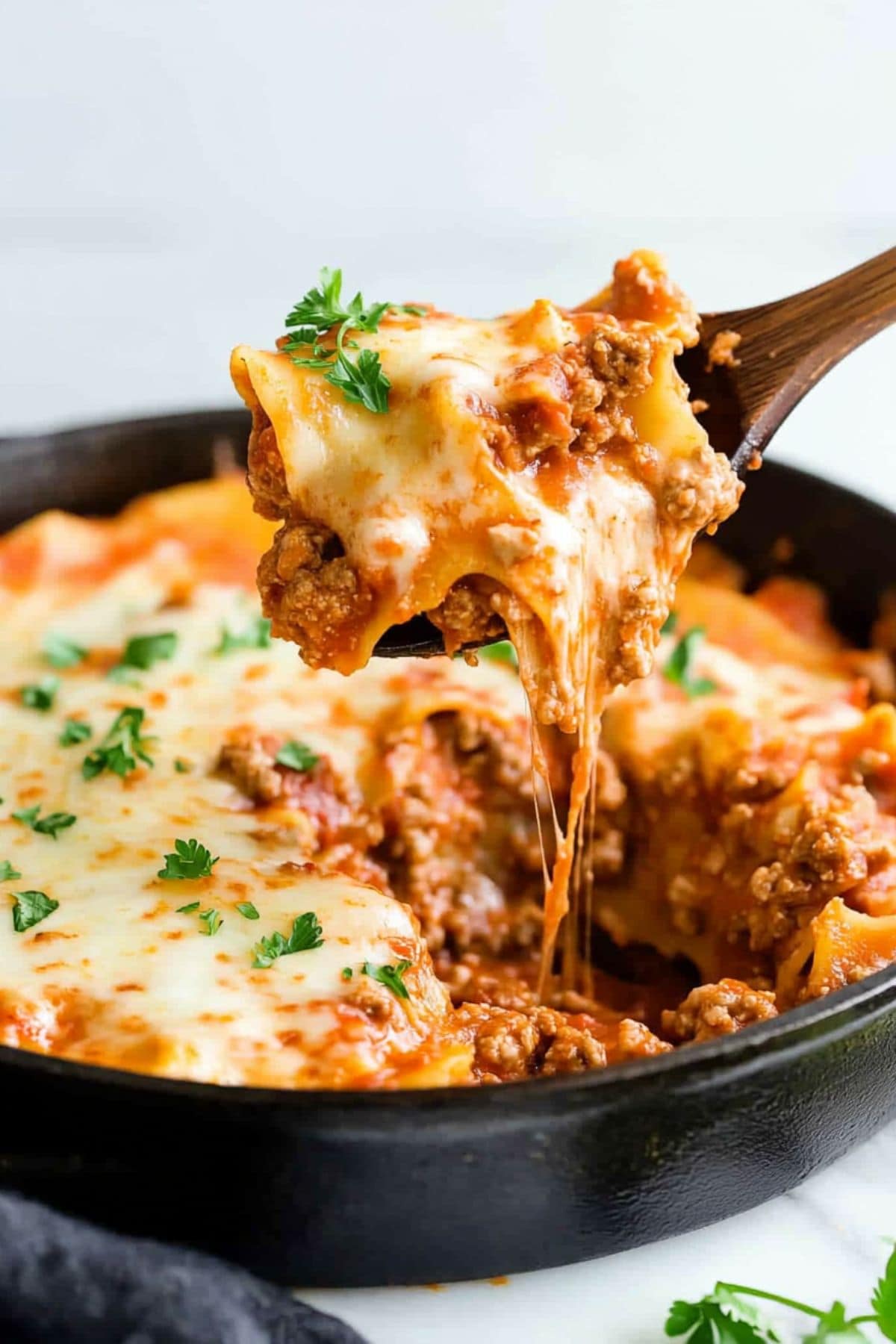 Wooden ladle scooping a serving of cheesy lasagna cooked in a skillet. 