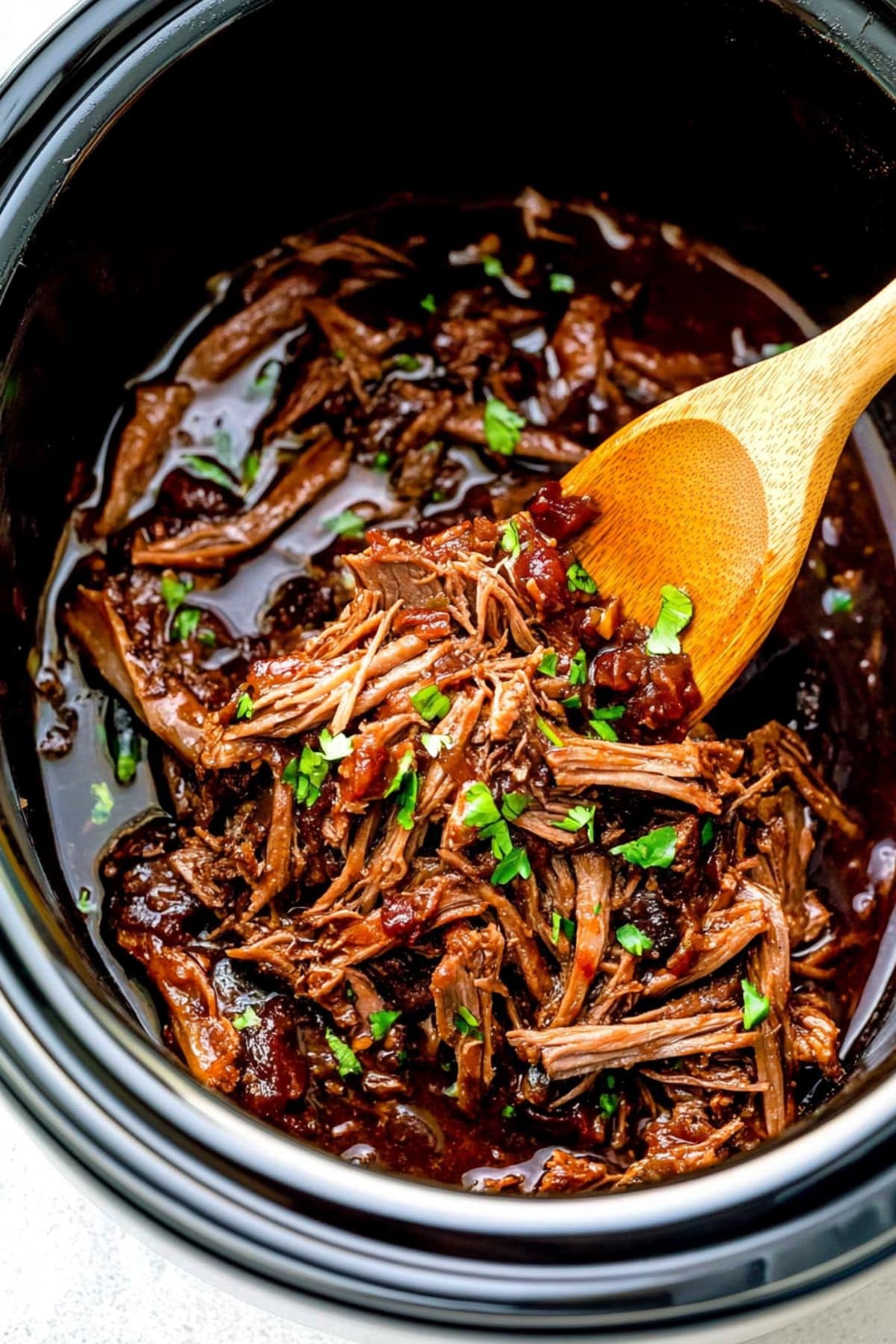 Pulled pork with Asian sauce inside a slow cooker with wooden spoon, close up, top view.