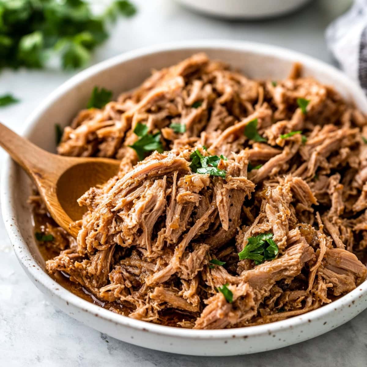 Wooden spoon scooping slow cooked shredded pork from a white bowl