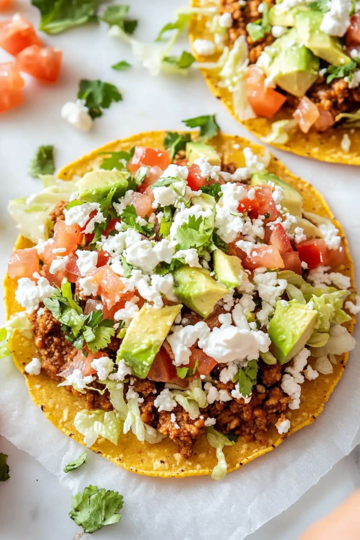 Tostadas topped with ground beef, chopped avocado, lettuce, crumbled cheese drizzled with sour cream sitting on white marble table.