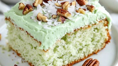 A square slice of fluffy and moist watergate cake with green frosting garnished with chopped pecans and shredded coconut served on a white plate.