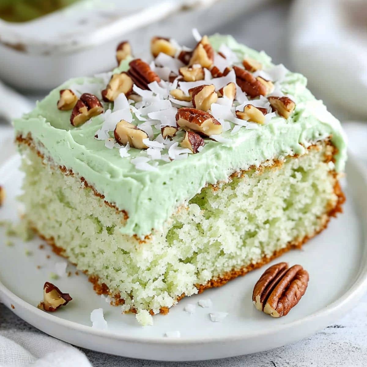 A square slice of fluffy and moist watergate cake with green frosting garnished with chopped pecans and shredded coconut served on a white plate.