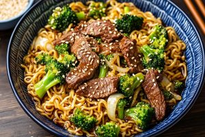 Beef and broccoli ramen stir fry served in a blue bowl garnished with sesame seeds.