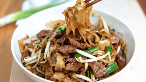 Wooden chopsticks picking noodles from a bowl serving of beef chow fun.