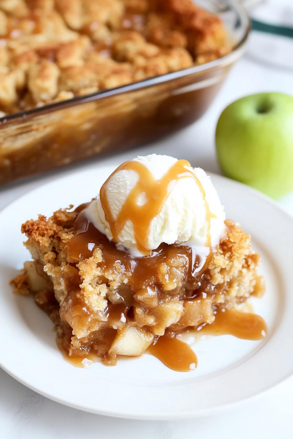 Serving of caramel apple dump cake on a white plate served with a scoop of vanilla ice cream and extra caramel sauce.