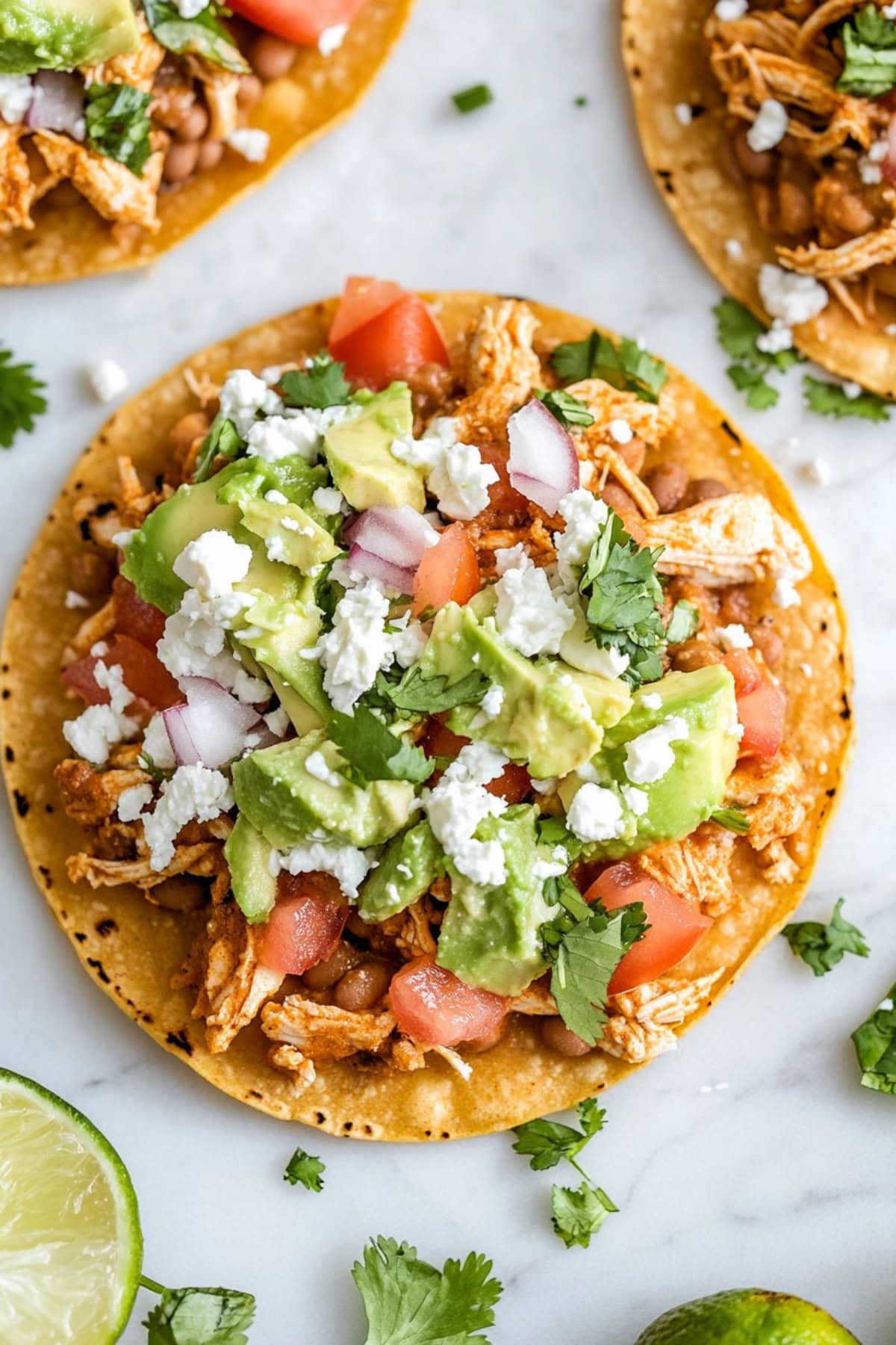 Chicken tostados made with Chicken tostados made with crispy corn tortilla topped with refried beans, mashed avocado, shredded chicken, lettuce, tomato, red onion, cotija cheese and cilantro flat lay on a marble table.
