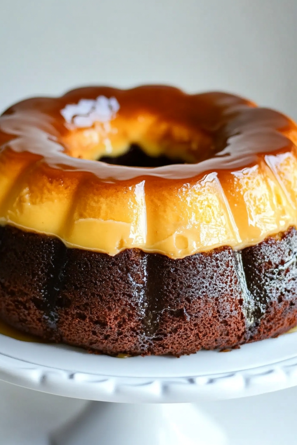 Moist chocolate cake topped with creamy flan sitting on a white cake stand, close up, side view. 